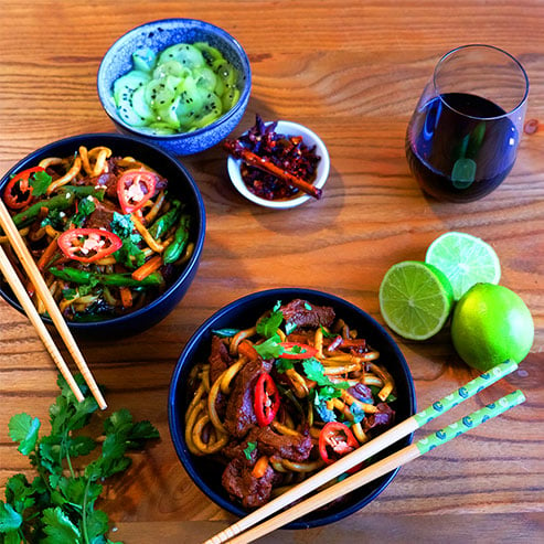 Two bowls of beef stir-fry noodles with chilli and limes and a glass of red wine