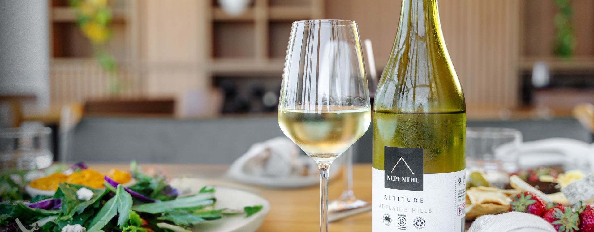 Nepenthe wine bottle and glass filled with white wine on a wooden table with salad in a bowl.