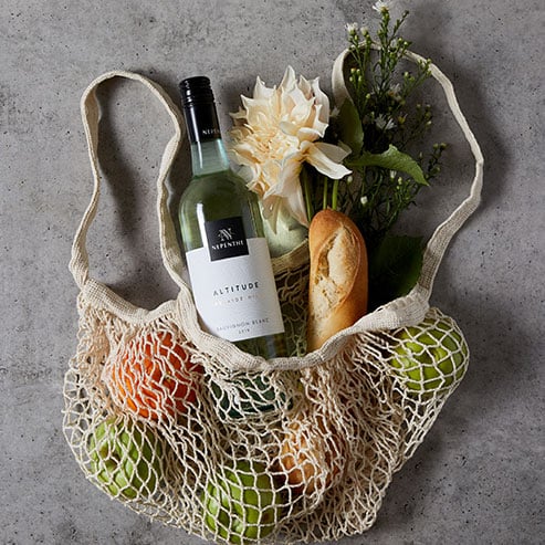 Bottle of Nepenthe Altitude Sauvignon Blanc in a crochet bag with a baguette and flowers against a black background