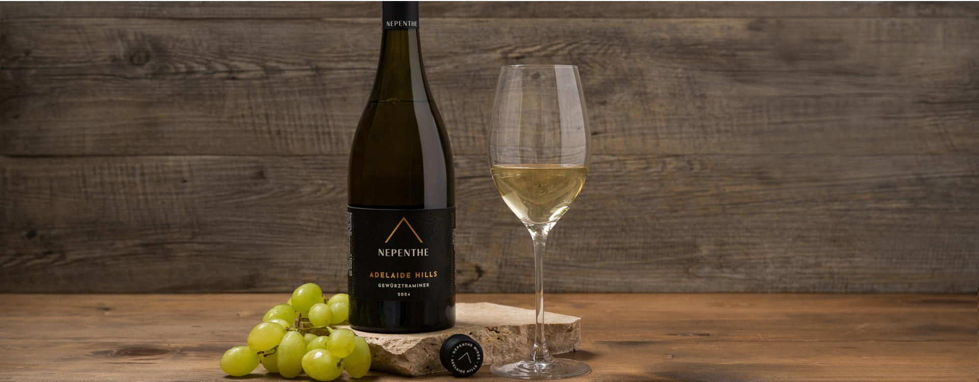 Nepenthe Pinnacle wine bottle with a poured glass bsdies. a bunch of grapes leaning in front of a wood rustic backdorp.